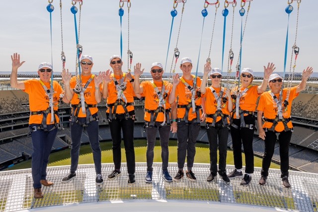 Picture of group with harnesses on waving at the camera and Optus Stadium in background.